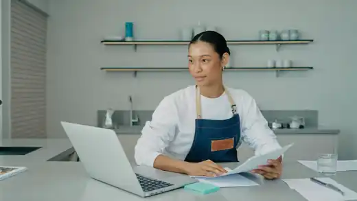 A small business owner at a bright shop counter reviewing finances on a laptop.